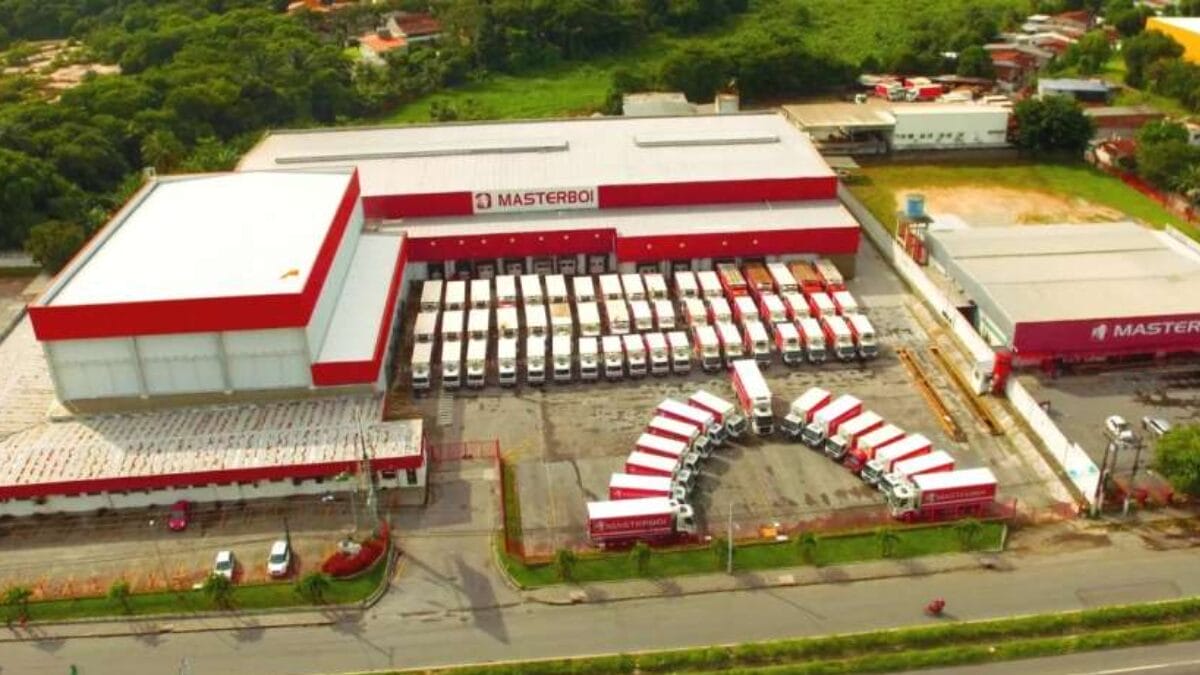 Vista a&eacute;rea de centro log&iacute;stico da Masterboi com dezenas de caminh&otilde;es estacionados em p&aacute;tio ao lado de galp&otilde;es industriais.