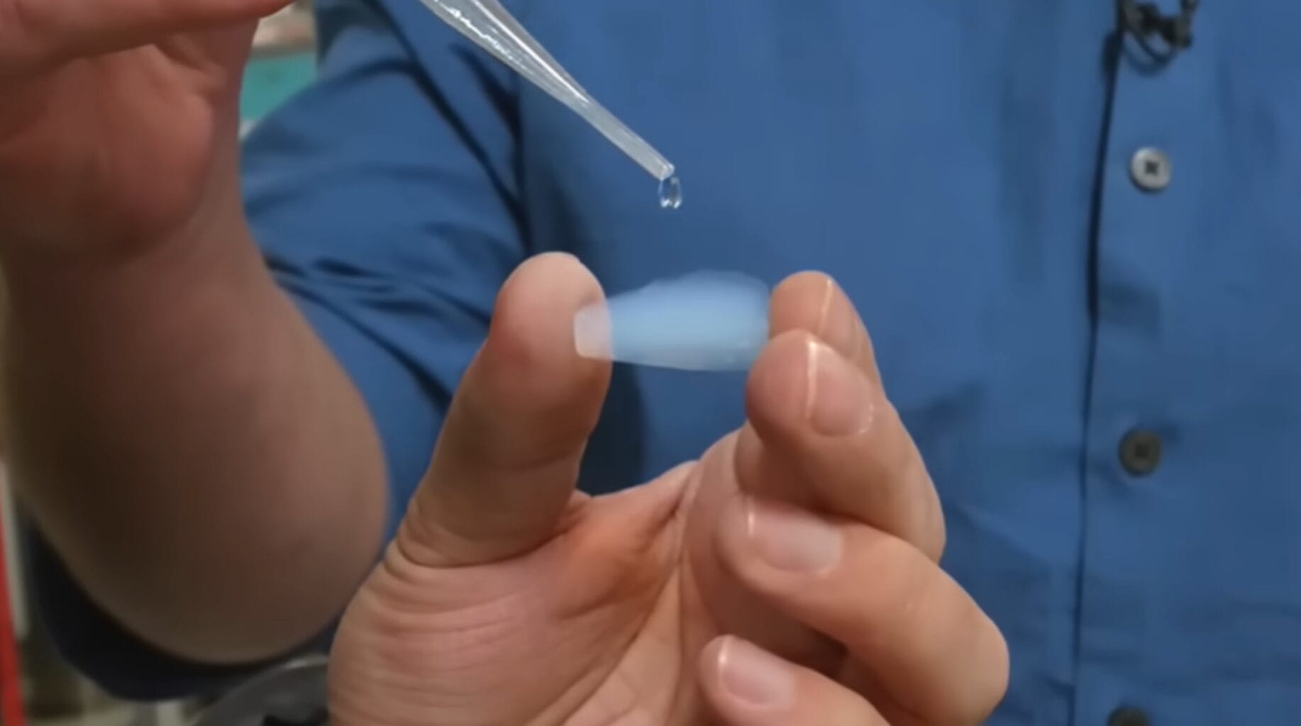 Gota de água caindo sobre pequena amostra de aerogel azul segurada entre os dedos