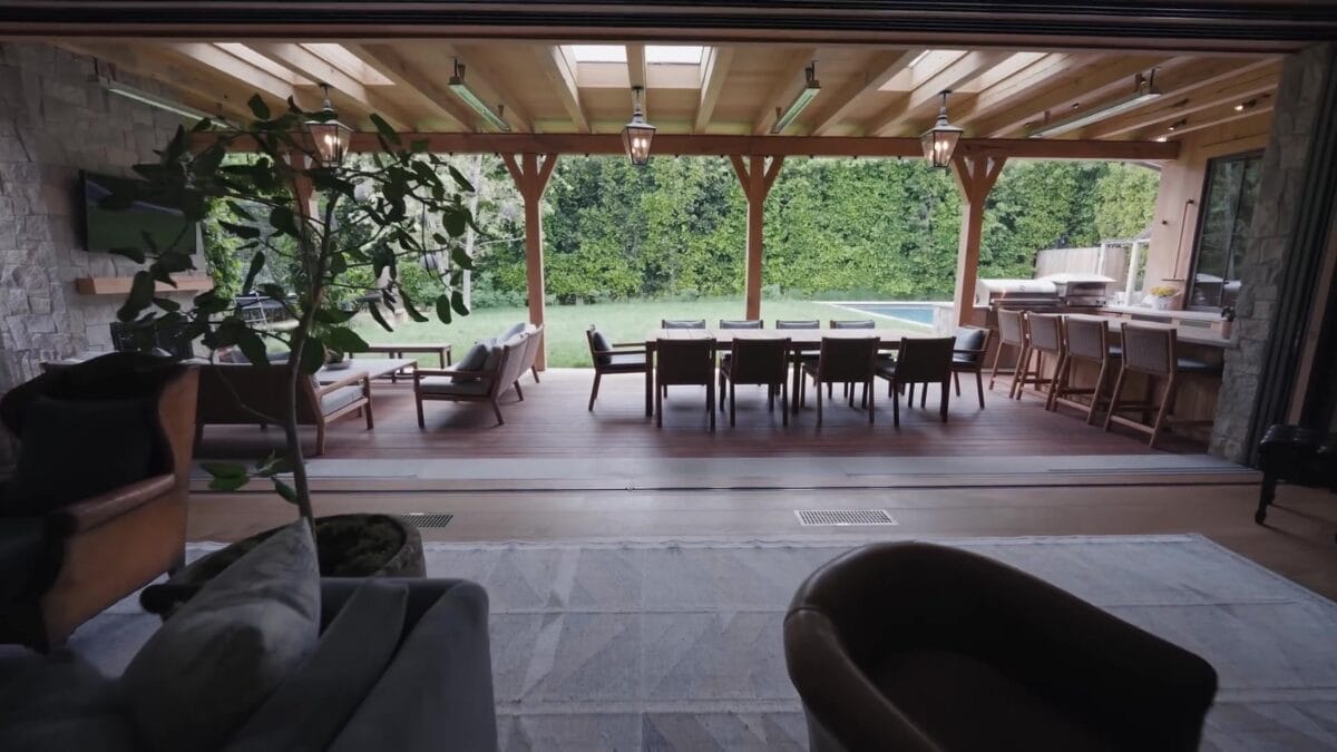  Sala ampla com piano de cauda preto &agrave; esquerda, vigas de madeira aparente no teto, parede de pedra natural e grandes portas abertas para o jardim.