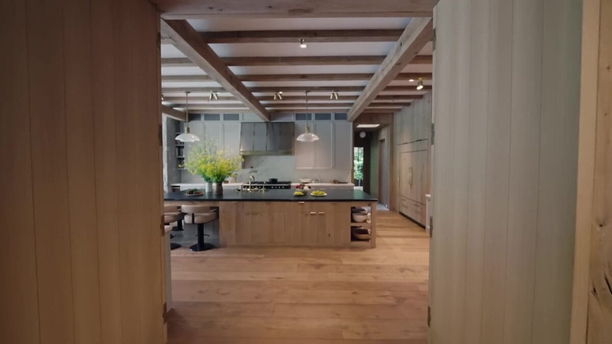 Cozinha ampla com grande ilha central em madeira clara e bancada escura, pendentes alinhados no teto e jardim vis&iacute;vel pelas janelas ao fundo.