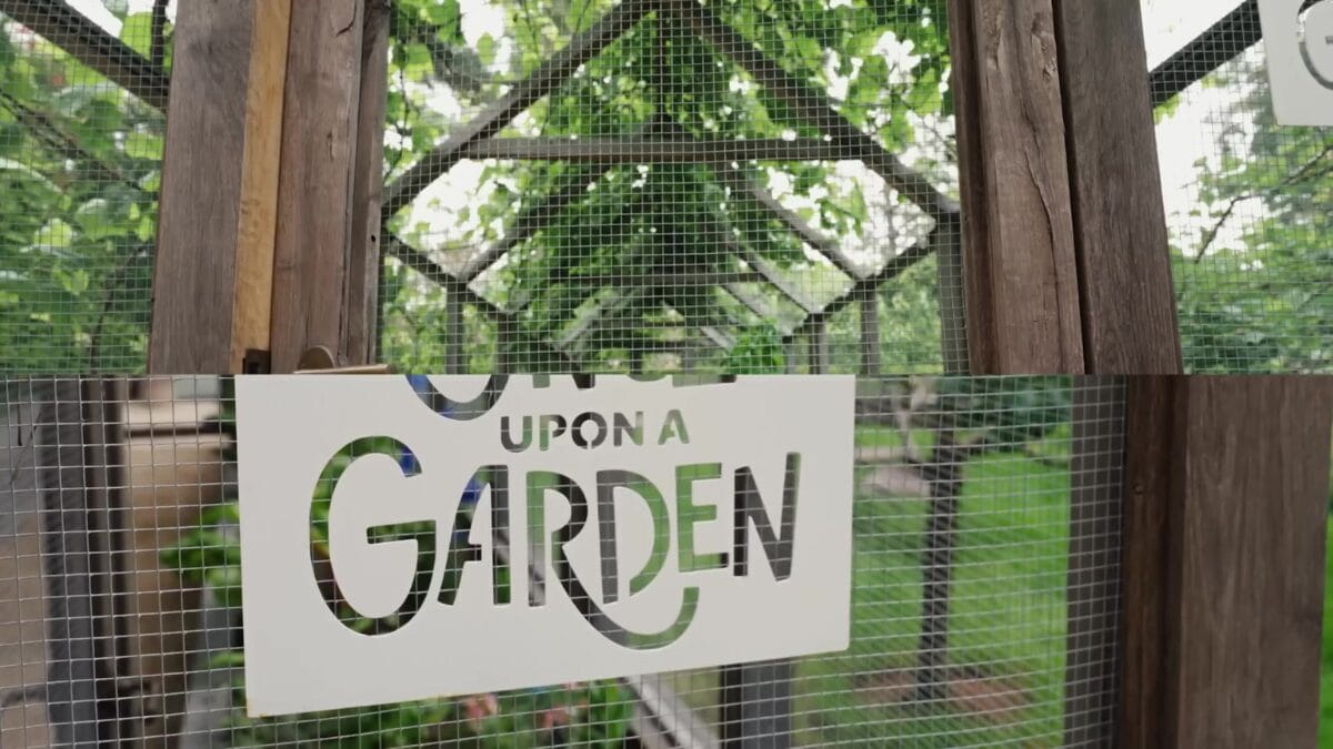 Entrada de estufa com estrutura de madeira e laterais teladas, exibindo placa pintada com a inscri&ccedil;&atilde;o "Upon a Garden" suspensa entre as plantas.