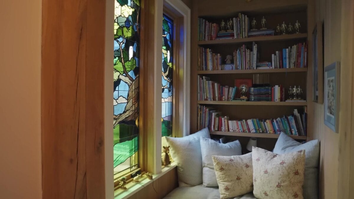 Cantinho de leitura com banco baixo coberto de almofadas estampadas, ladeado por estantes de livros infantis e iluminado por vitrais coloridos com motivos de natureza.
