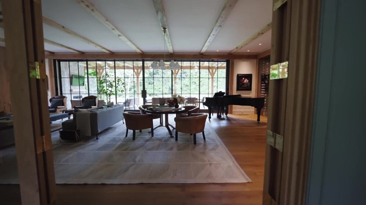 Sala de estar e jantar vistas atrav&eacute;s de portas duplas de madeira, com tapete claro, poltronas, mesa de centro e piano ao fundo.