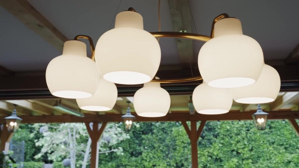 Pendente de teto com seis globos brancos de vidro suspensos por hastes de lat&atilde;o, com sala de estar e janelas voltadas para o jardim ao fundo.