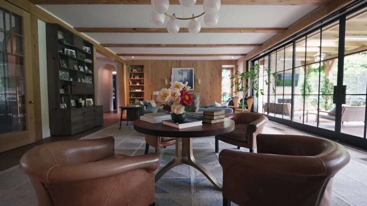 Lounge com poltronas de couro marrom em torno de mesa redonda de madeira escura com arranjos florais, e parede envidra&ccedil;ada &agrave; direita exibindo adega climatizada.