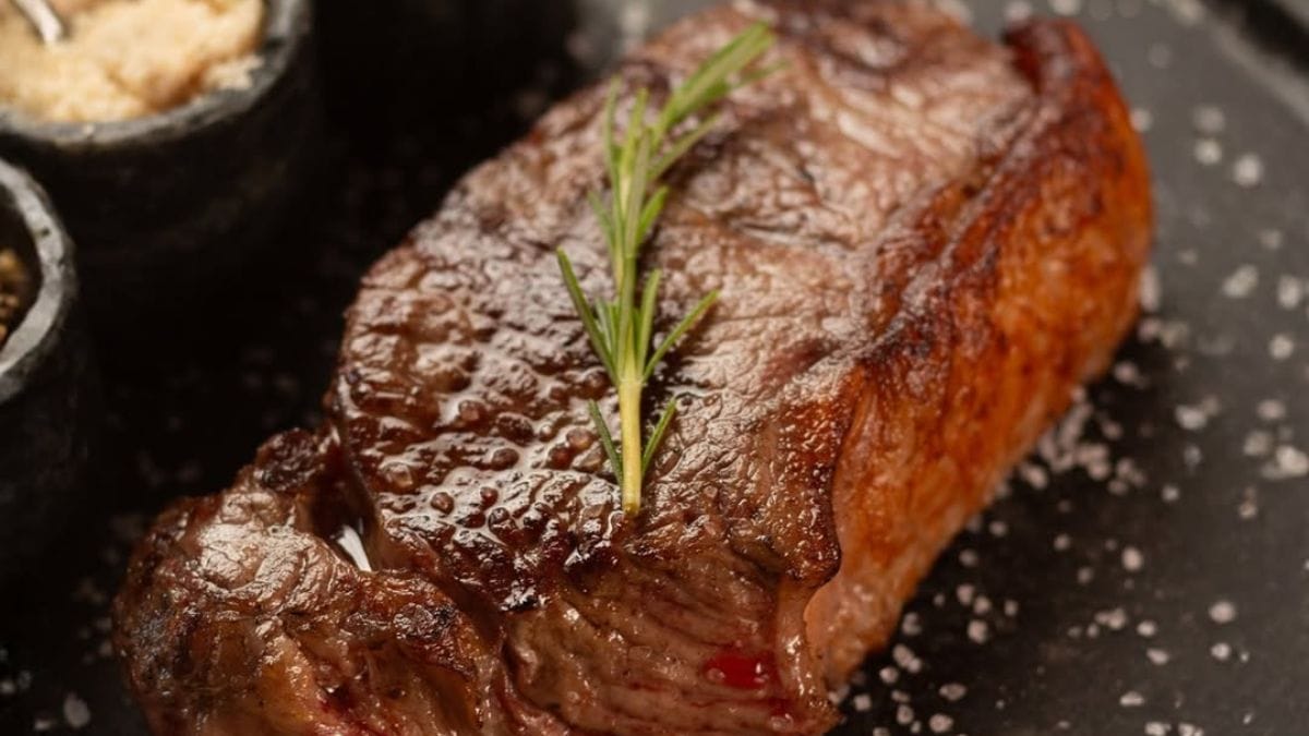 Pedaço de carne bovina assada, com ramo de alecrim por cima, disposta sobre suprfície na cor preta com grãos de sal grosso, do Los Troncos Parrilla Uruguaya.