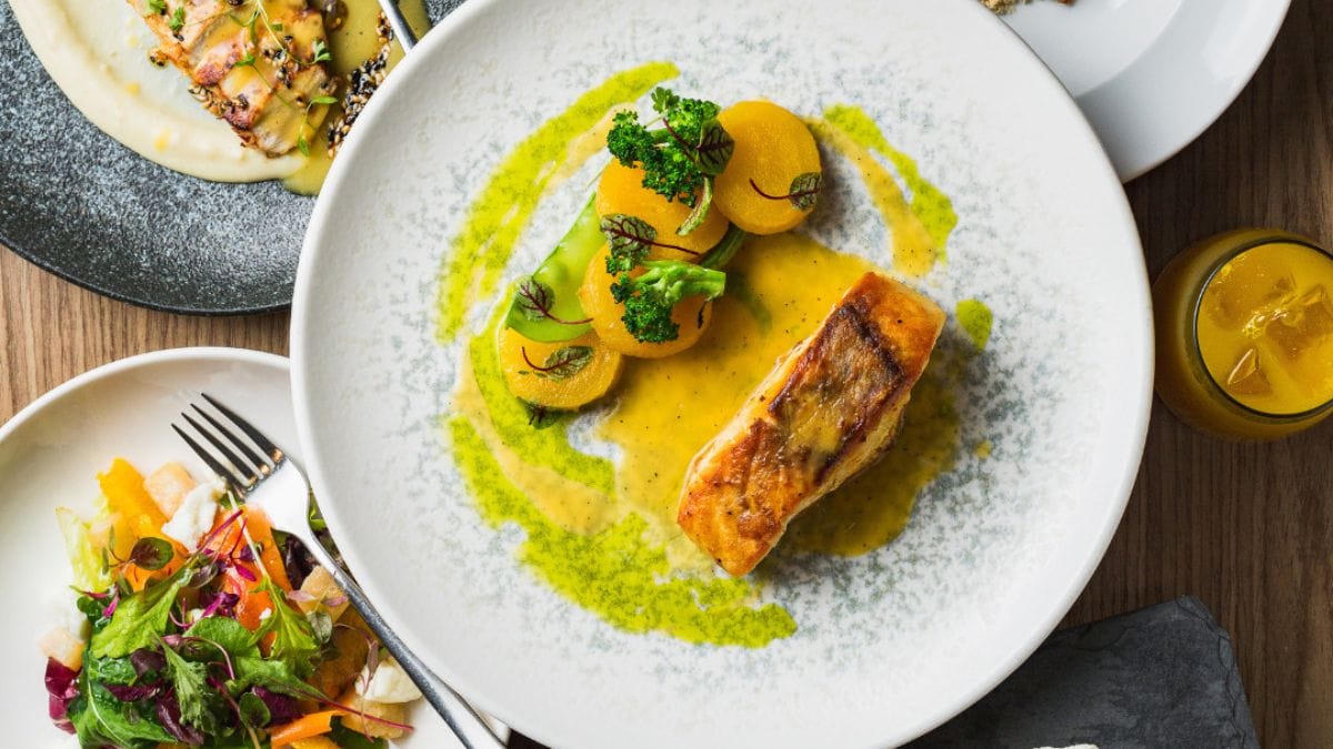 Prato com pedaço de salmão acompanhado de rodelas de legumes e ervas e molho na cor amarelo, do Osli Restaurante.