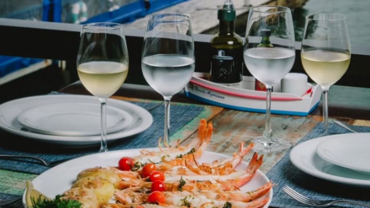 Mesa posta com taças de vinho servidas e prato generoso com frutos do mar. Ao fundo se vê parte da água do mar, do Rancho Açoriano.