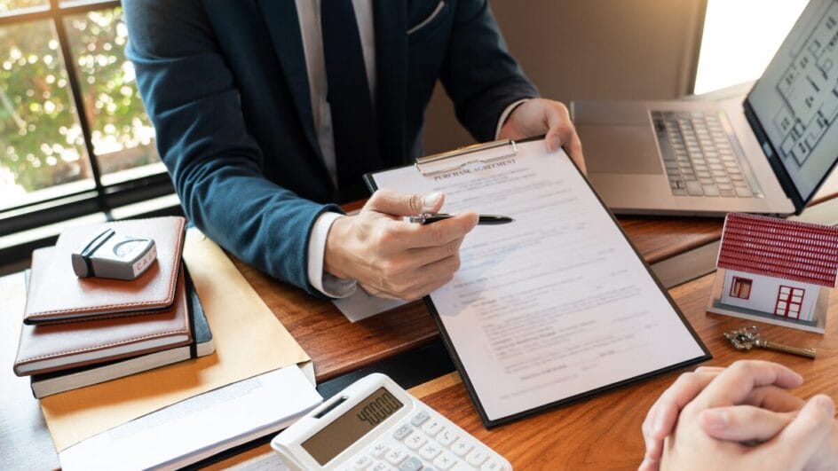 pessoa de terno mostra contrato para assinatura em uma mesa com calculadora e notebook