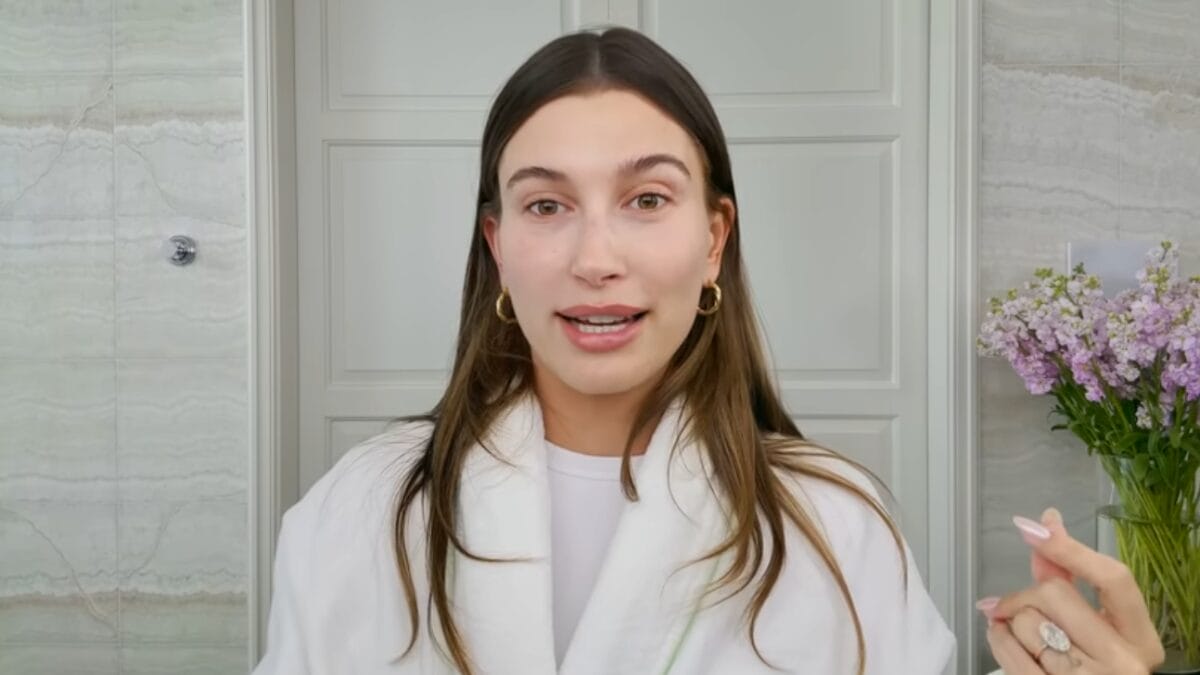 Hailey Bieber fala para a c&acirc;mera usando roup&atilde;o branco durante v&iacute;deo de skincare minimalista.