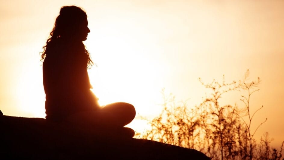 Silhueta de mulher meditando ao pôr do sol em ambiente ao ar livre.