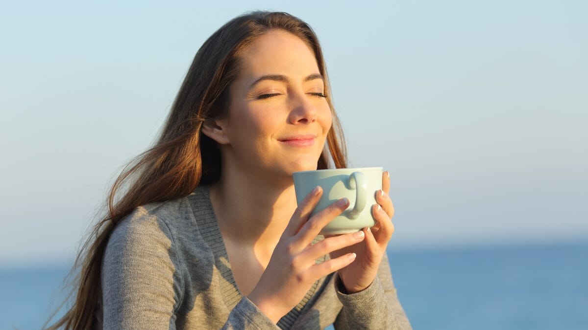 Mulher segurando uma caneca ao ar livre com os olhos fechados, aproveitando a luz do sol.
