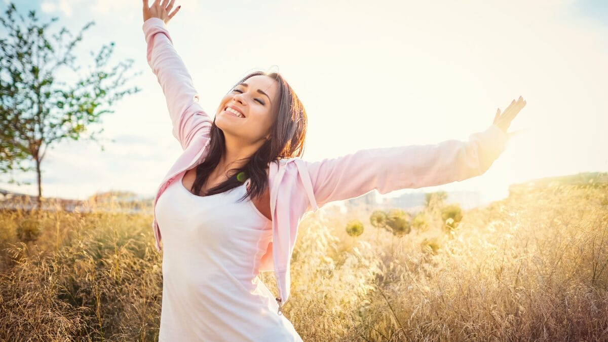 Mulher sorrindo com os braços abertos em campo iluminado pela luz do sol.