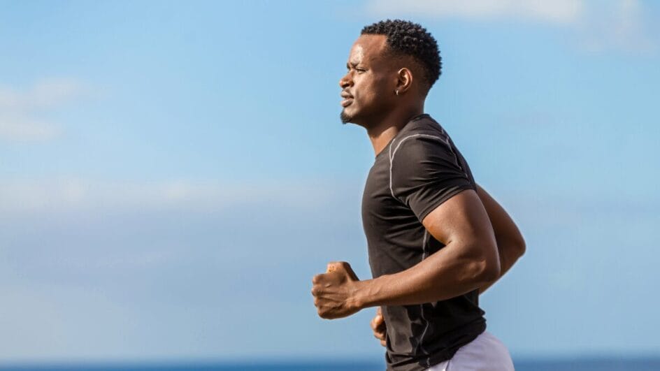 Homem praticando corrida ao ar livre próximo ao mar, vestindo camiseta esportiva preta.