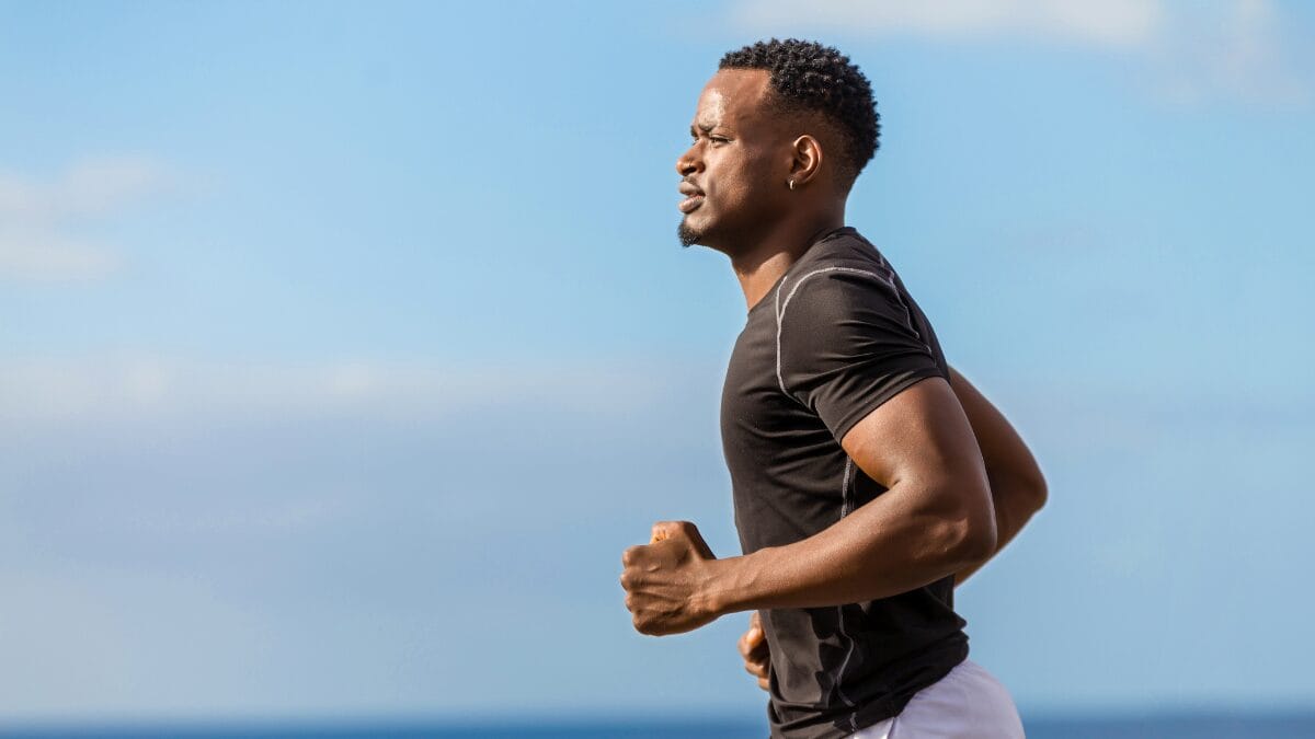 Homem praticando corrida ao ar livre próximo ao mar, vestindo camiseta esportiva preta.