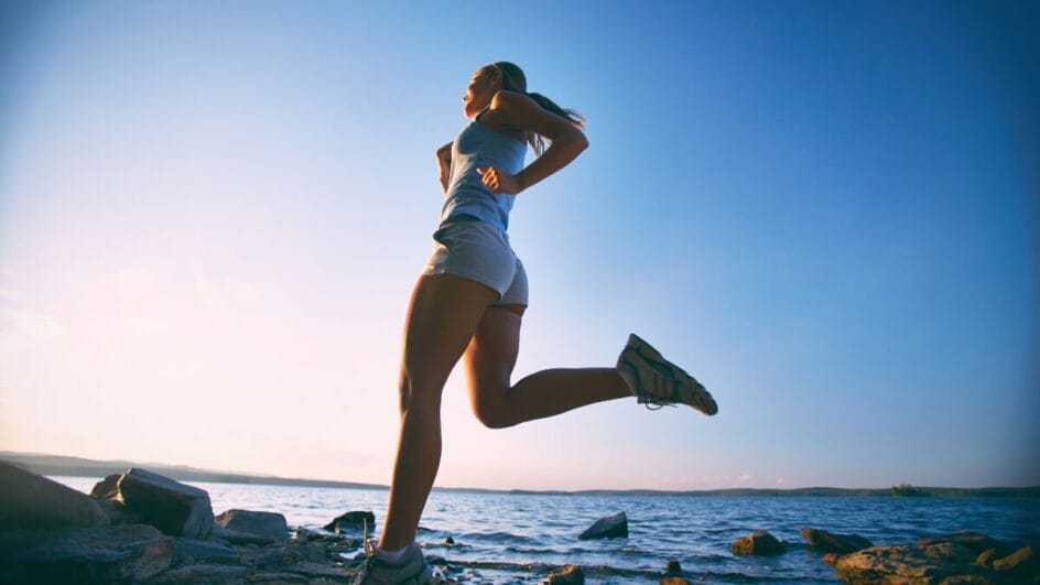Mulher correndo à beira de um lago durante o pôr do sol, usando roupas esportivas claras e tênis, em cenário ao ar livre.