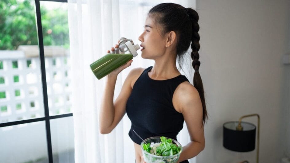 Mulher com roupa esportiva segurando uma salada e bebendo suco verde próximo à janela de casa.