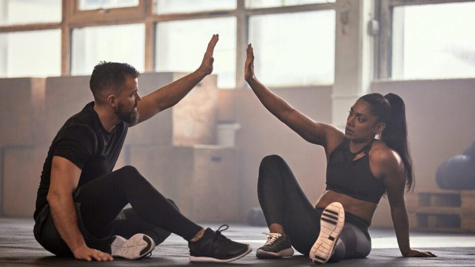 Homem e mulher sentados no chão de academia fazendo cumprimento com as mãos após treino físico.
