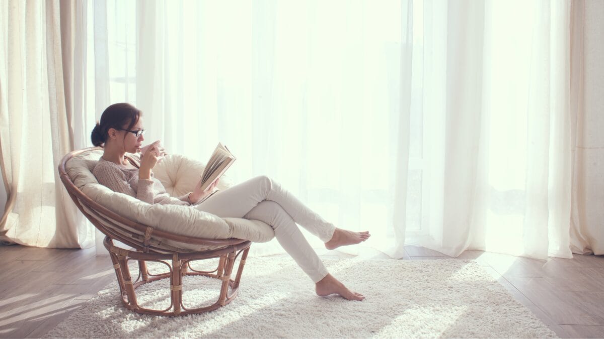 Mulher sentada em poltrona lendo um livro e tomando chá em ambiente iluminado pela luz natural.