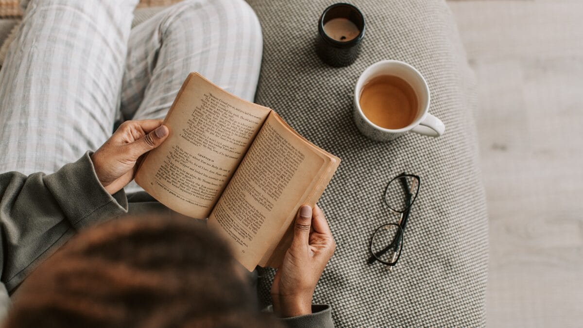Pessoa lendo livro no sofá ao lado de uma xícara de chá e óculos sobre a mesa.