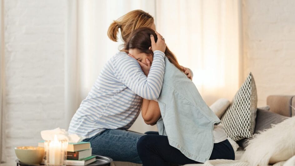 Mulher abraçando outra pessoa em gesto de apoio emocional dentro de casa.