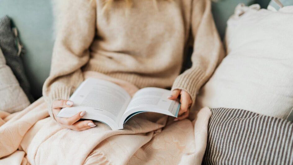 Pessoa enrolada em manta lendo livro no sofá, usando roupas de inverno em ambiente aconchegante.