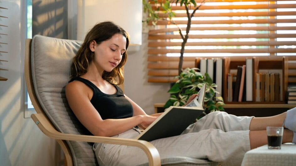Mulher relaxando em poltrona enquanto lê um livro em sala aconchegante com plantas ao fundo.