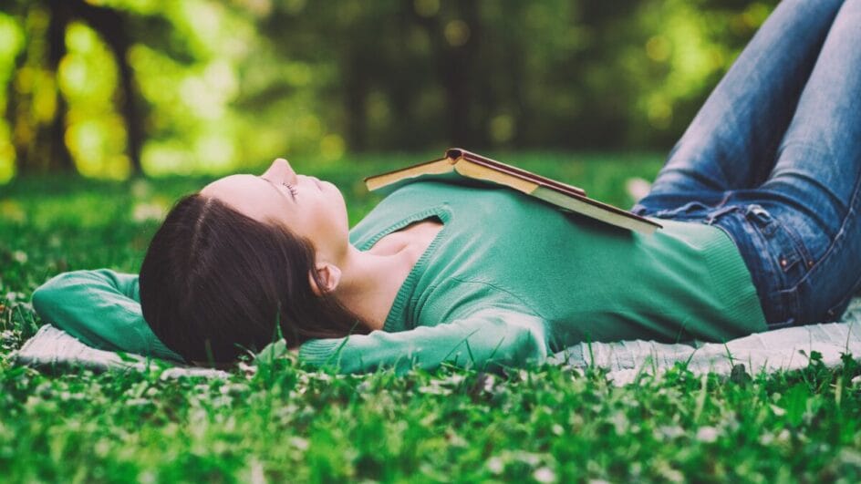 Mulher deitada na grama com um livro sobre o peito em momento de descanso ao ar livre.