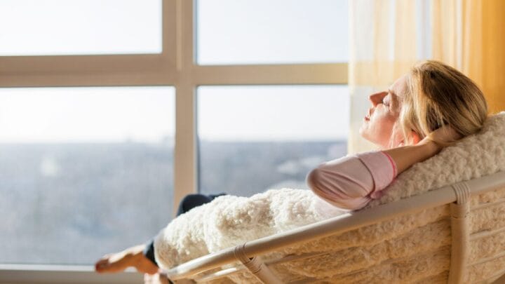 Mulher relaxando em cadeira próxima à janela, coberta com manta e aproveitando a luz do sol.