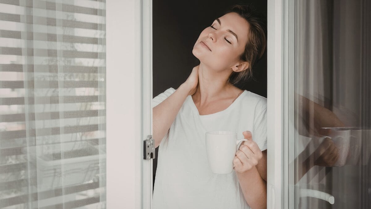 Mulher segurando uma caneca enquanto respira profundamente na janela de casa durante a manhã.