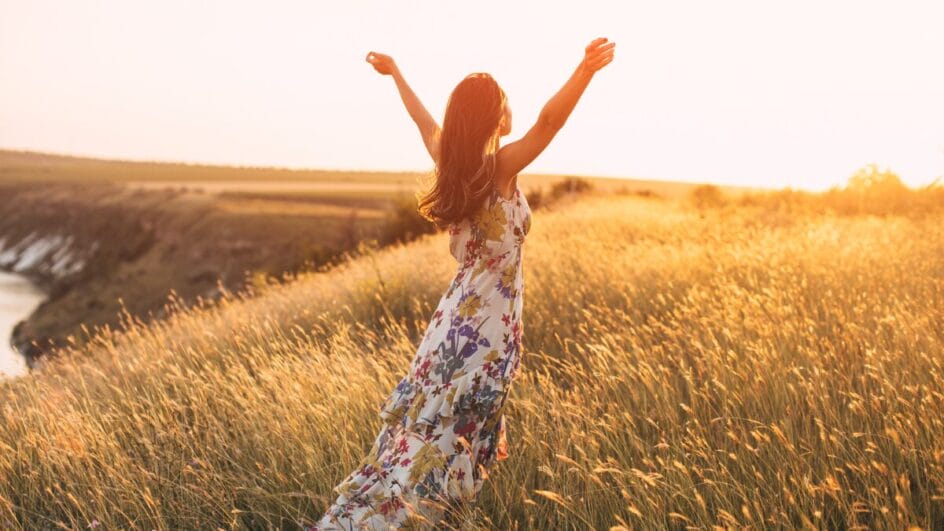 Mulher caminhando em campo dourado ao pôr do sol, usando vestido floral e braços erguidos.