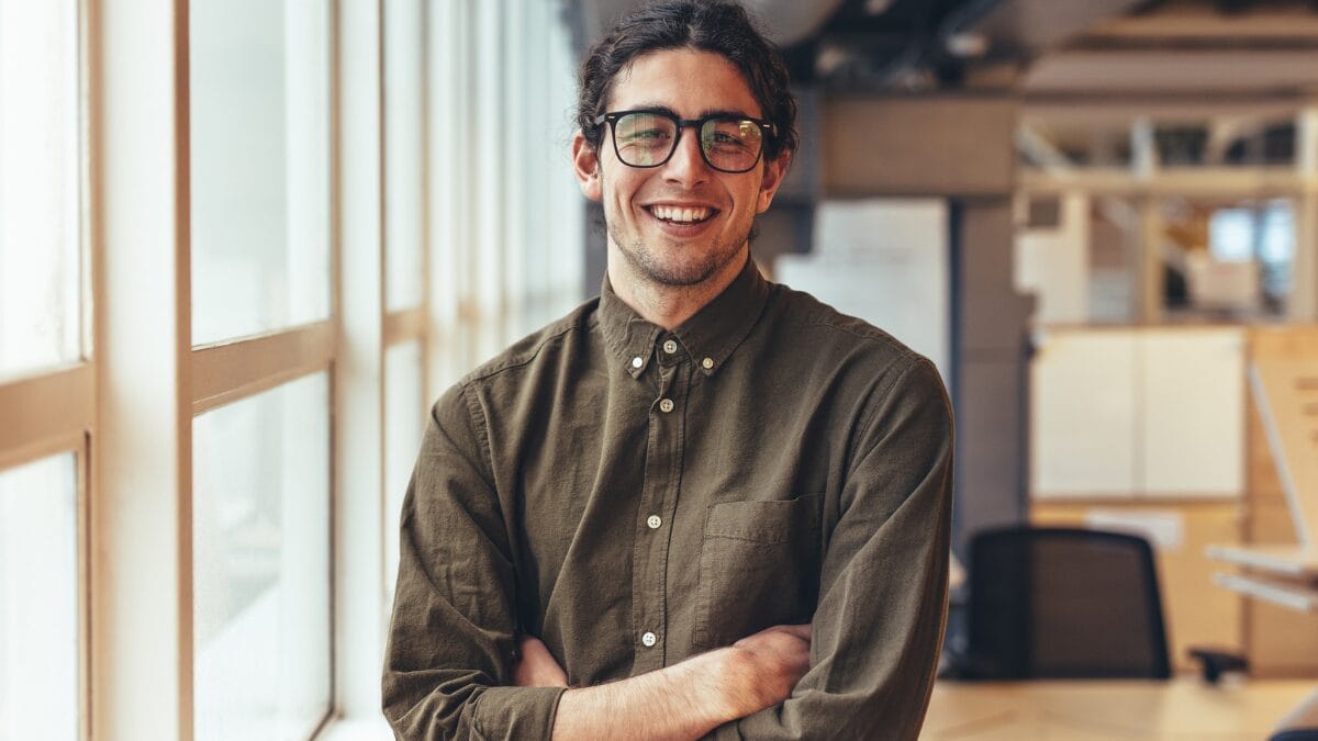 Homem sorrindo com os braços cruzados em ambiente de escritório iluminado.