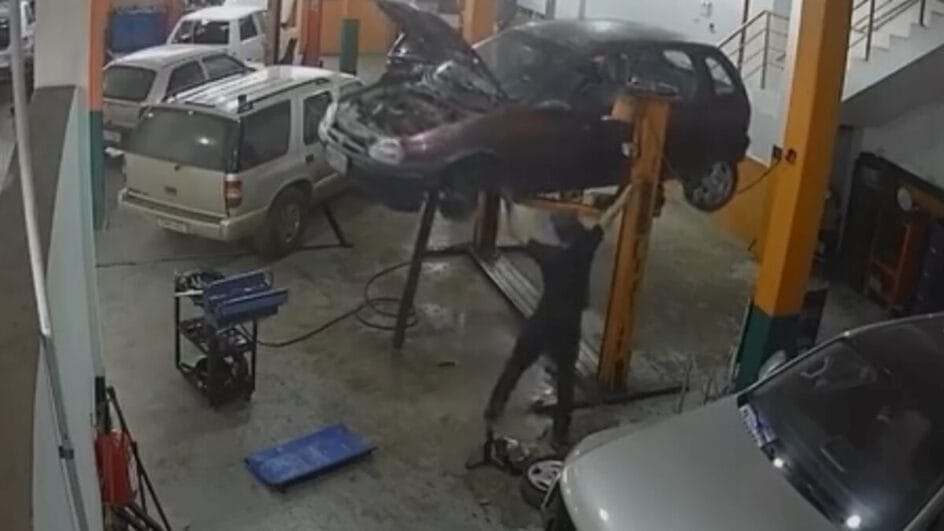 imagem de câmera de segurança que mostra um carro de cor escura suspenso por um elevador automotivo operado por um homem de roupas escuras