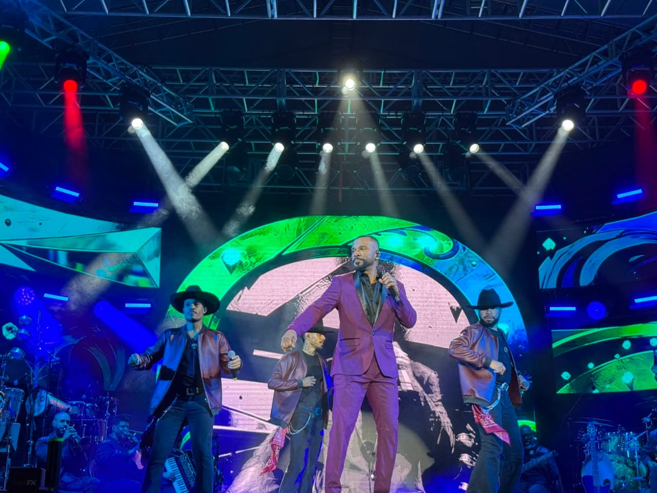 Close de Alexandre Pires no palco, cantando com terno roxo sob ilumina&ccedil;&atilde;o azul, com a banda e dan&ccedil;arinos desfocados ao fundo.