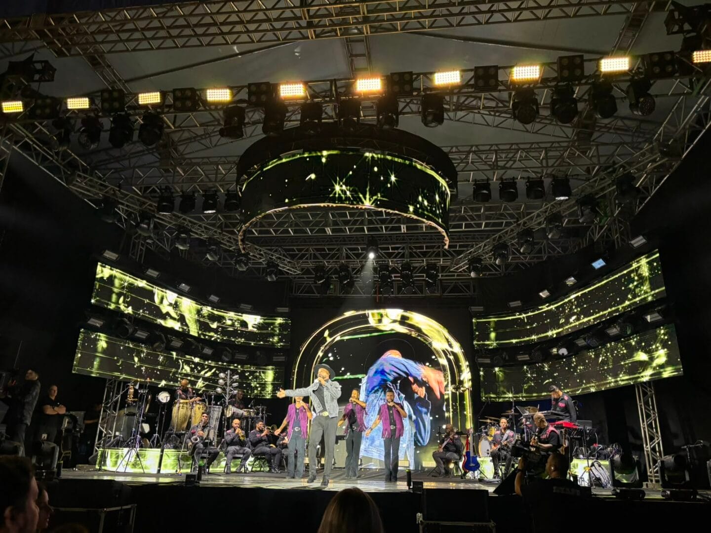 Close de Alexandre Pires no palco, cantando com terno roxo sob ilumina&ccedil;&atilde;o azul, com a banda e dan&ccedil;arinos desfocados ao fundo.
