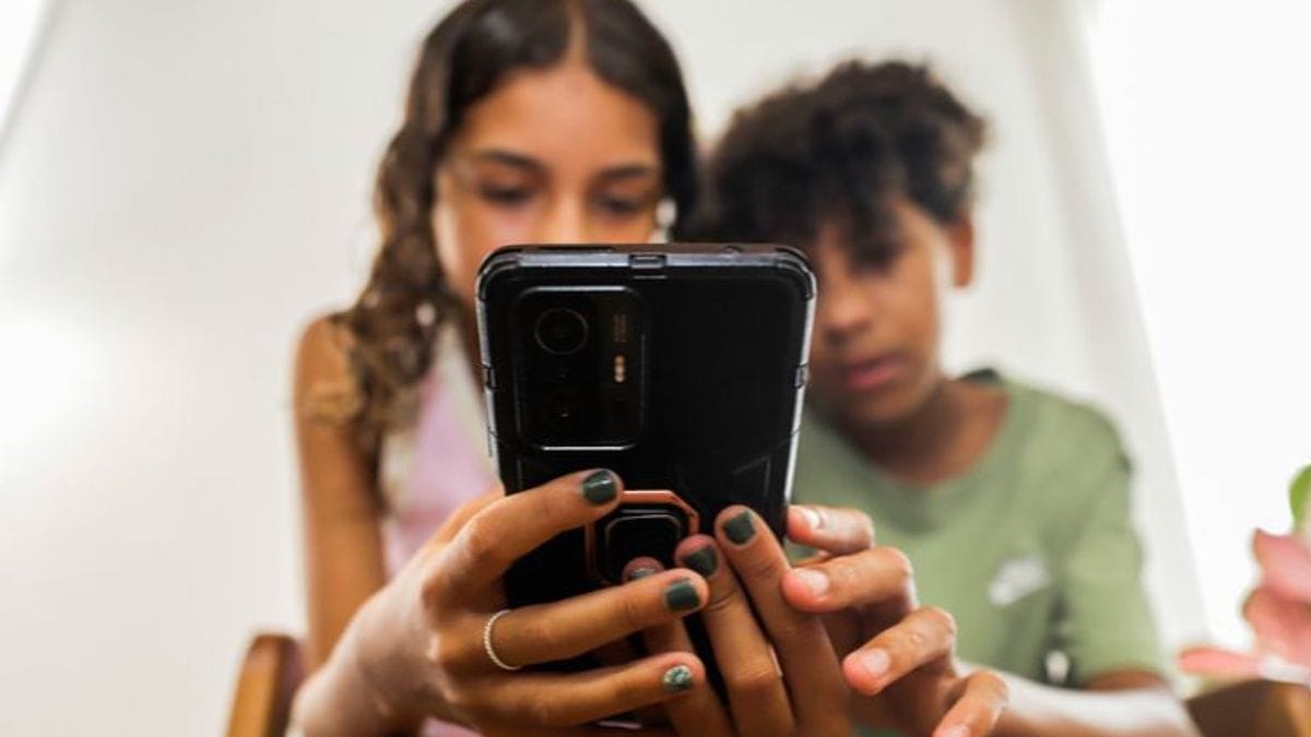 Foto mostra duas crianças sentadas olhando ao mesmo tempo para um celular que está em uma das mãos de ambos