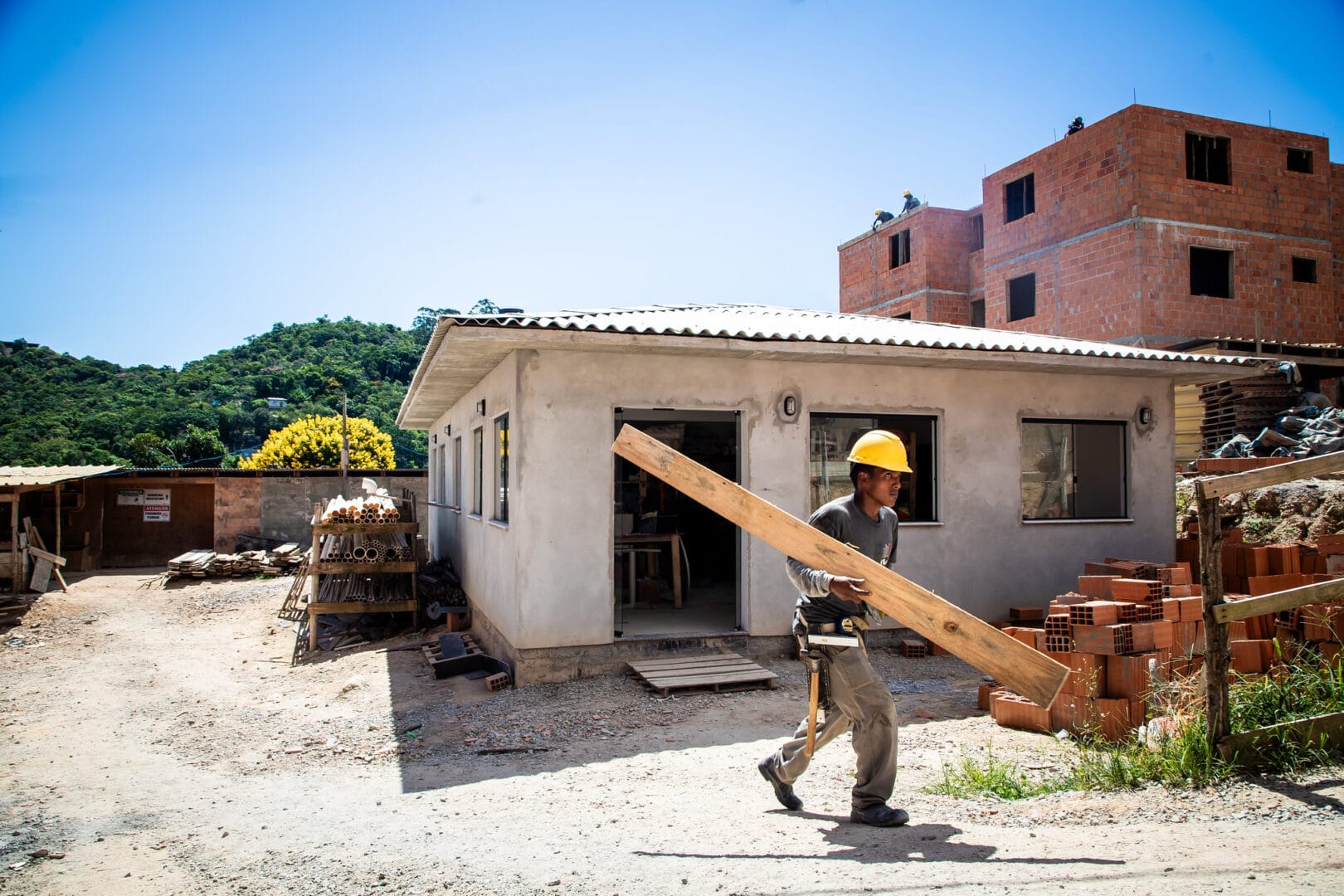Operário de capacete amarelo caminha em um canteiro de obras carregando uma grande viga de madeira no ombro diante de prédios de tijolos em construção.