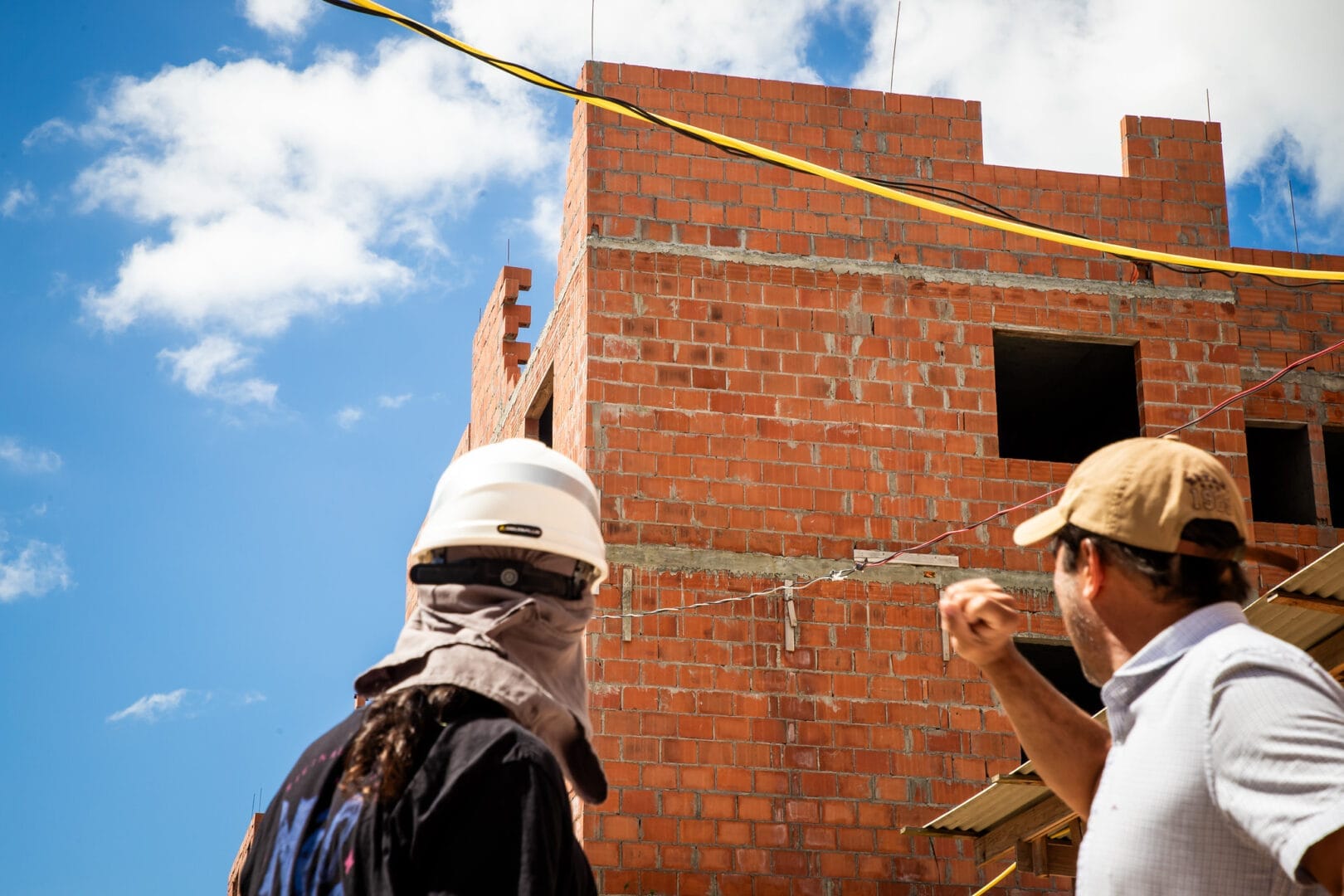Dois engenheiros ou mestres de obras de capacete observam e apontam em direção ao topo de um edifício de tijolos à vista em construção.