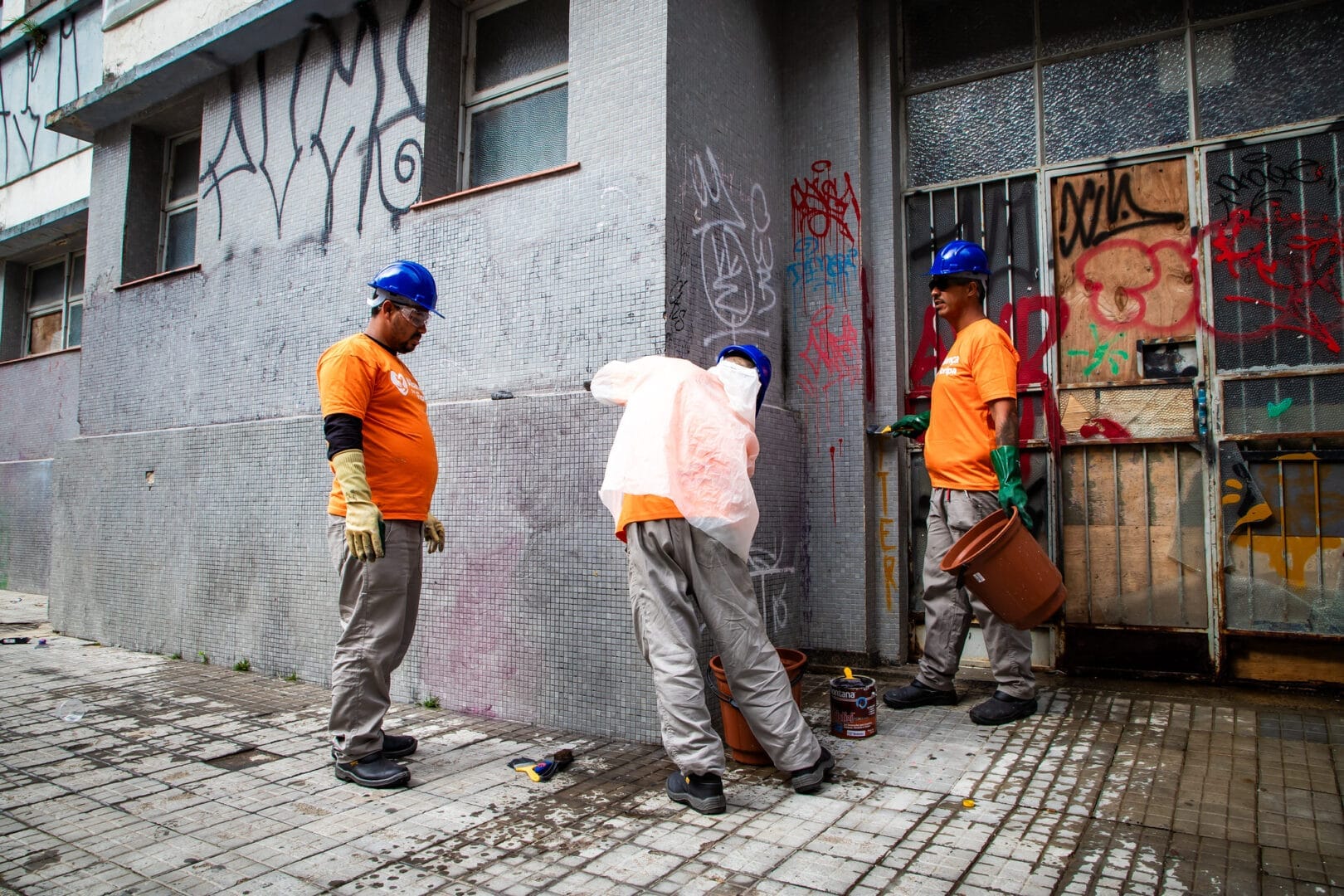 Três operários de capacete azul e uniforme realizam manutenção e pintura na fachada cinza pastilhada de um prédio antigo com pichações.