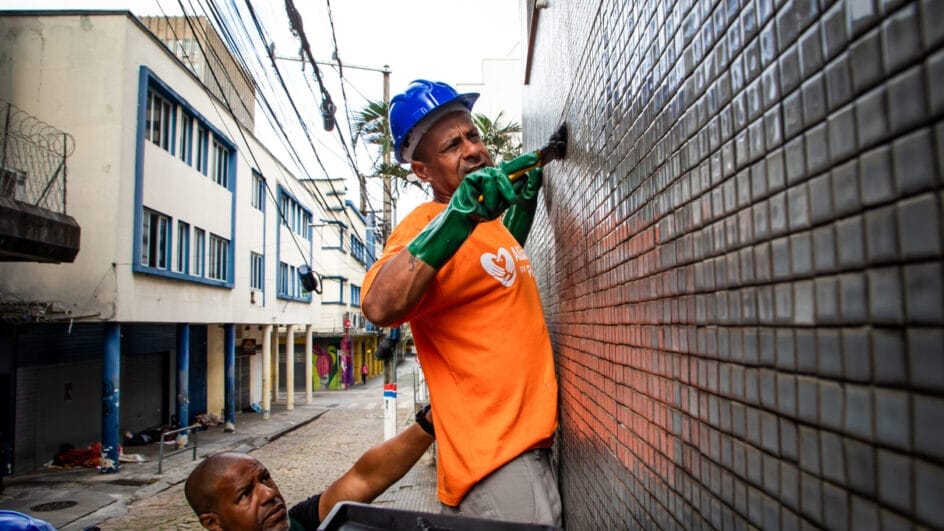 Operário com luvas verdes e camisa laranja pinta a parede pastilhada de uma rua estreita enquanto outro homem segura a bandeja de tinta abaixo.