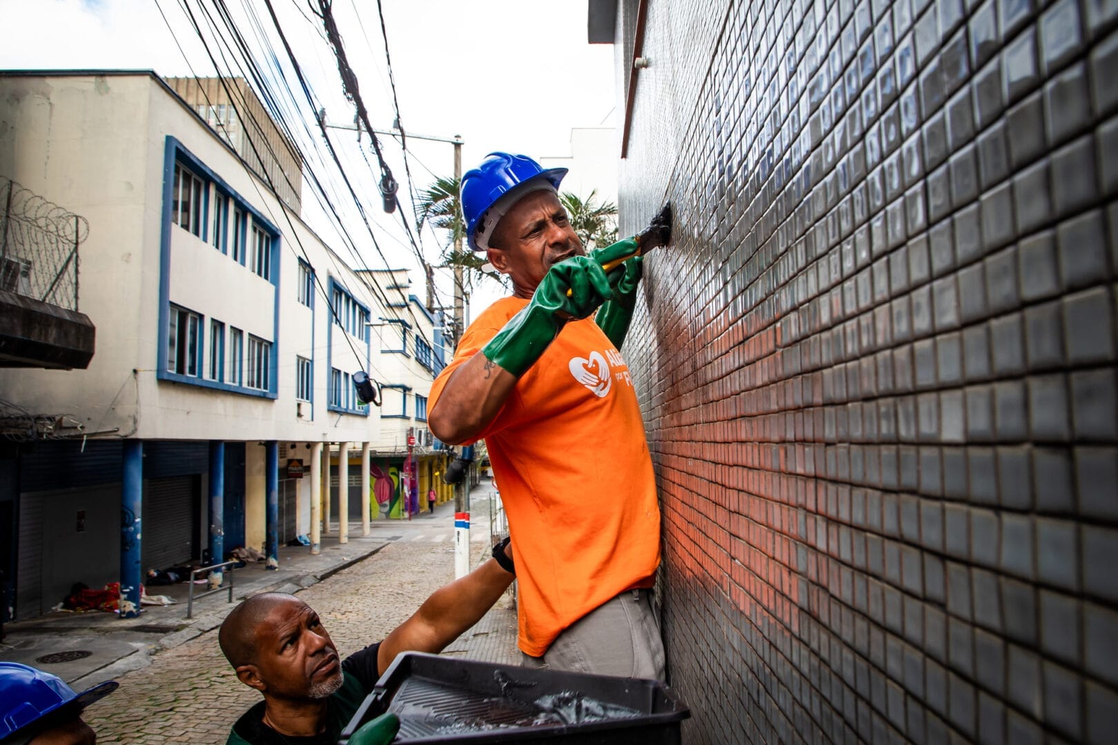Operário com luvas verdes e camisa laranja pinta a parede pastilhada de uma rua estreita enquanto outro homem segura a bandeja de tinta abaixo.