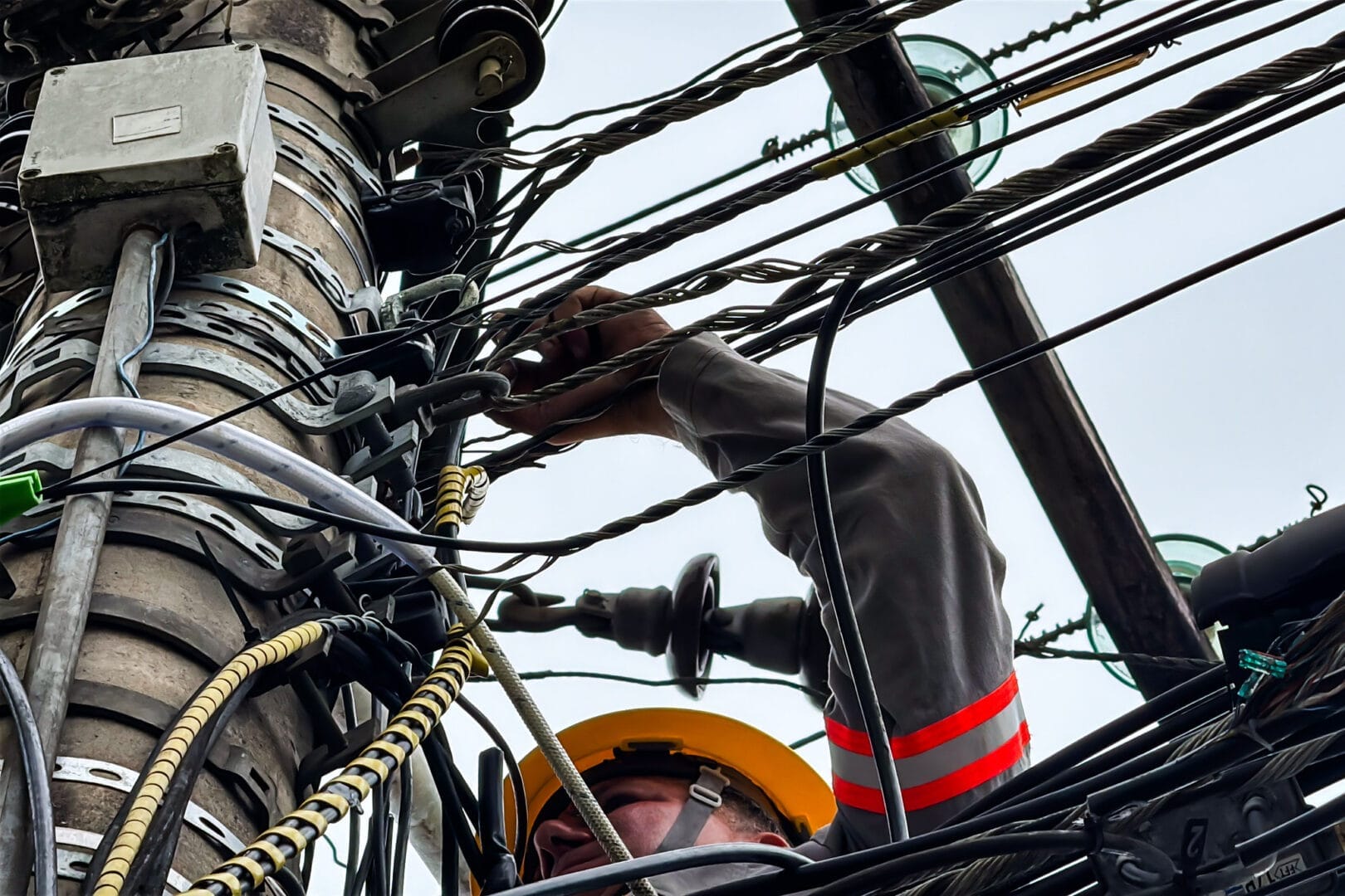 Vista de baixo para cima de um eletricista de capacete amarelo trabalhando em meio a um emaranhado de fios em um poste de energia elétrica.