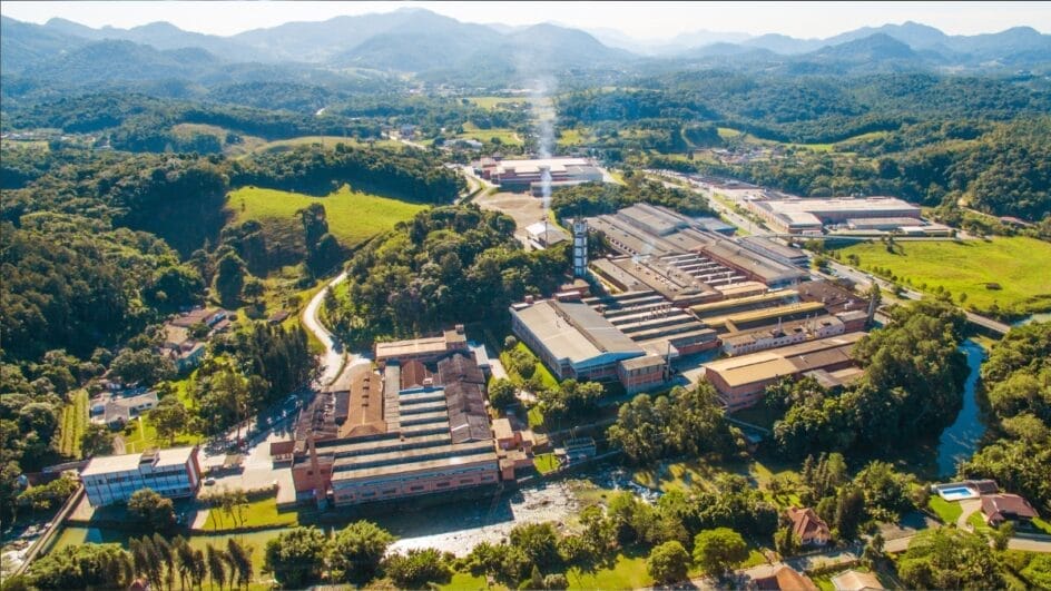 Vista aérea do parque fabril da Karsten no bairro Testo Salto, em Blumenau, com galpões industriais cercados por morros verdes e vegetação nativa