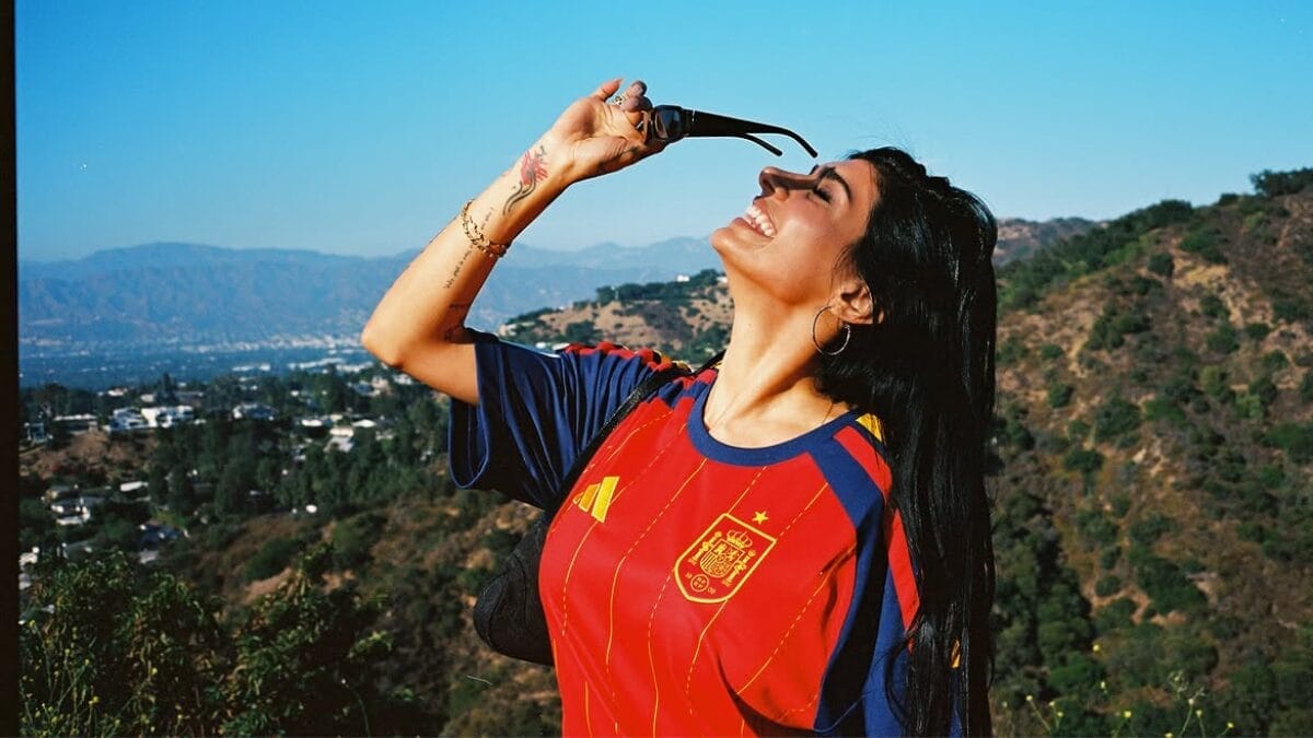 Mia Khalifa sorri enquanto ergue um par de óculos escuros acima do rosto, usando uma camisa vermelha da seleção da Espanha, em um mirante com montanhas ao fundo.