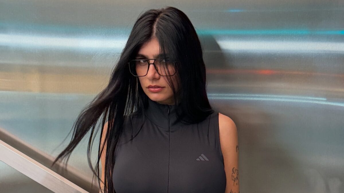 Mia Khalifa posa com expressão séria diante de uma parede metálica, usando óculos de armação preta e blusa preta sem mangas.
