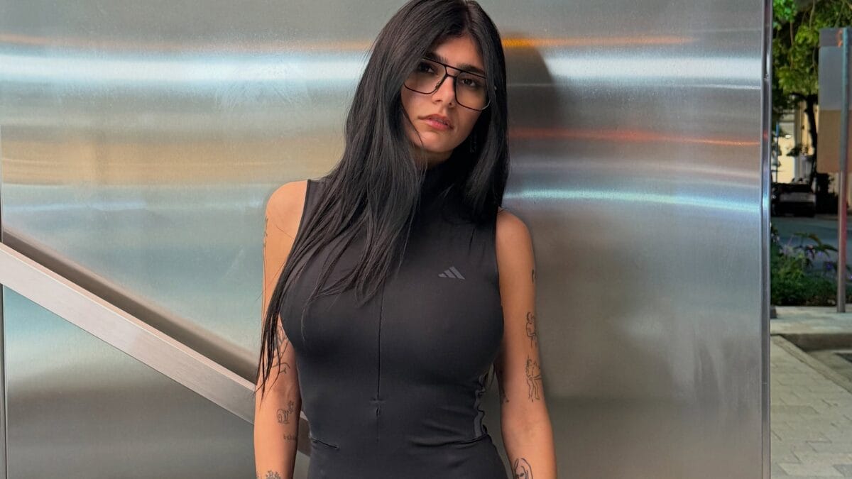 Mia Khalifa posa diante de uma parede metálica usando óculos de armação preta e blusa preta de gola alta sem mangas.