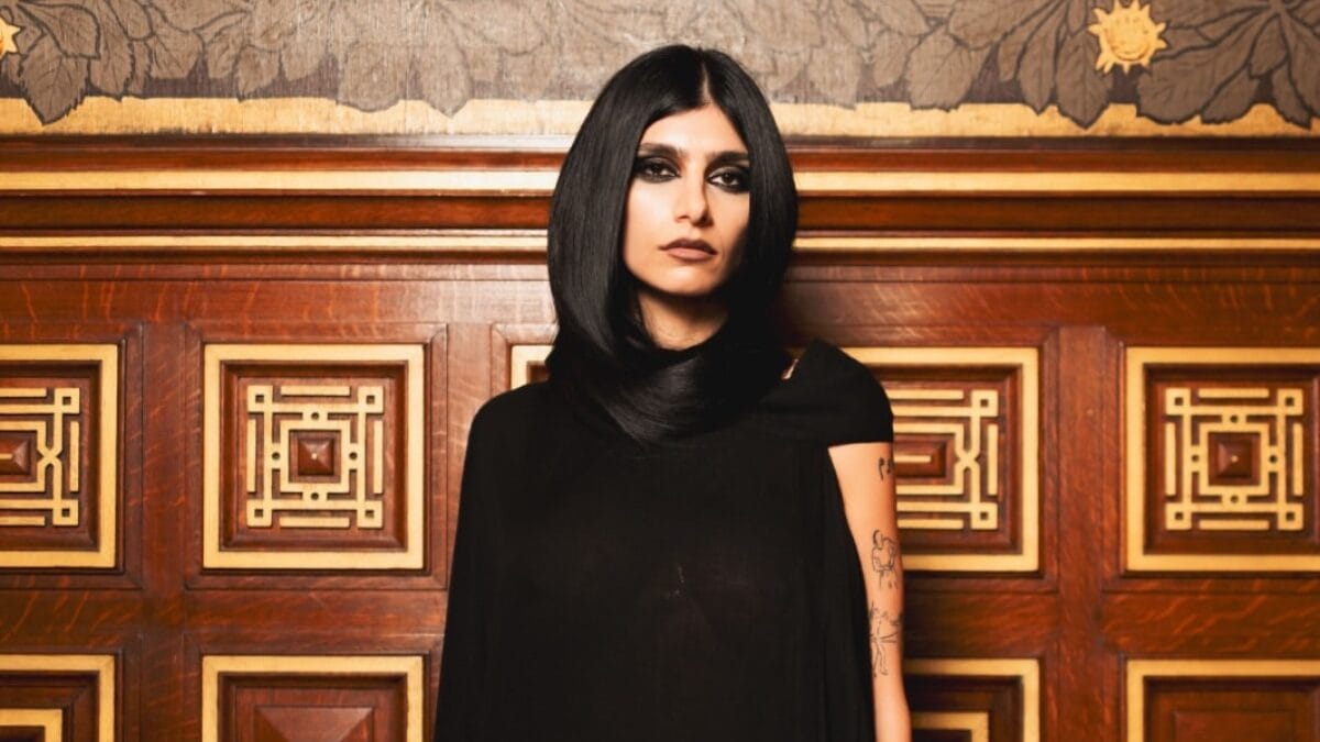 Mia Khalifa posa com maquiagem escura e roupa preta diante de um painel de madeira ornamentado.