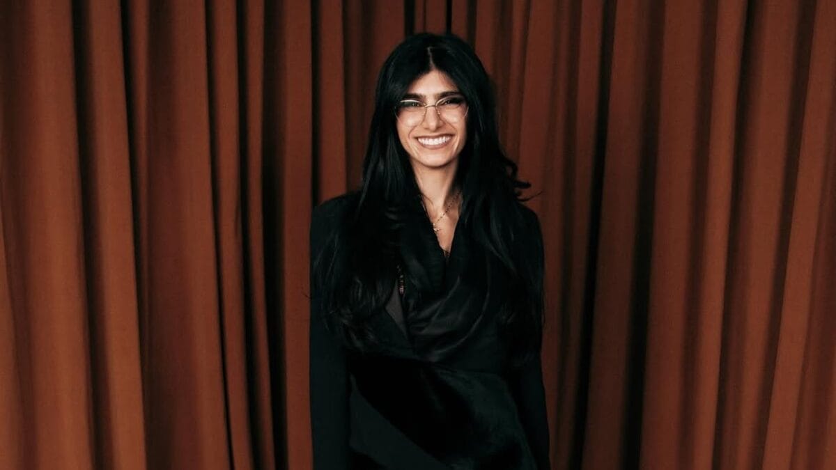 Mia Khalifa sorri para a câmera usando óculos de armação fina e roupa preta diante de uma cortina marrom.