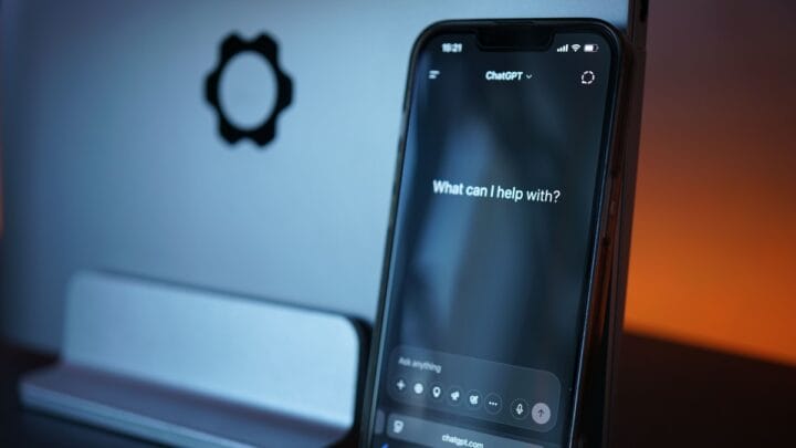 Tela de um iPhone exibindo o aplicativo do ChatGPT ao lado de um MacBook de alumínio sobre uma mesa de escritório escura.