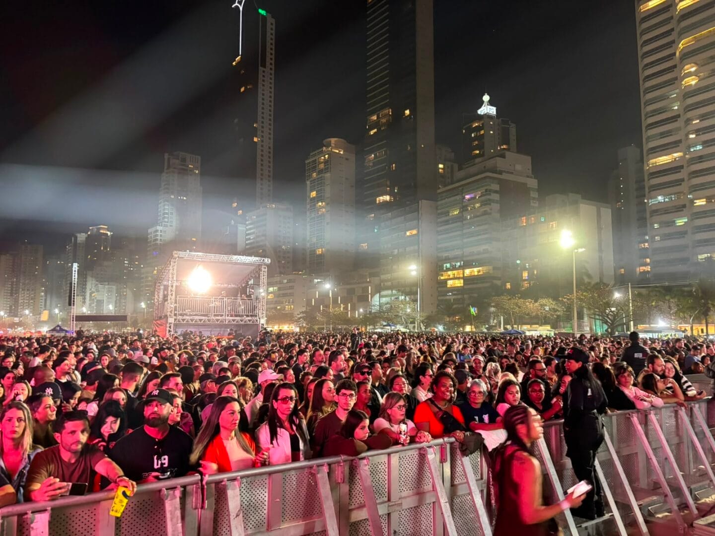 Vis&atilde;o panor&acirc;mica de uma multid&atilde;o de pessoas reunida na Praia Central de Balne&aacute;rio Cambori&uacute; &agrave; noite, aguardando o show, de frente para um palco iluminado, com arranha-c&eacute;us da cidade ao fundo.